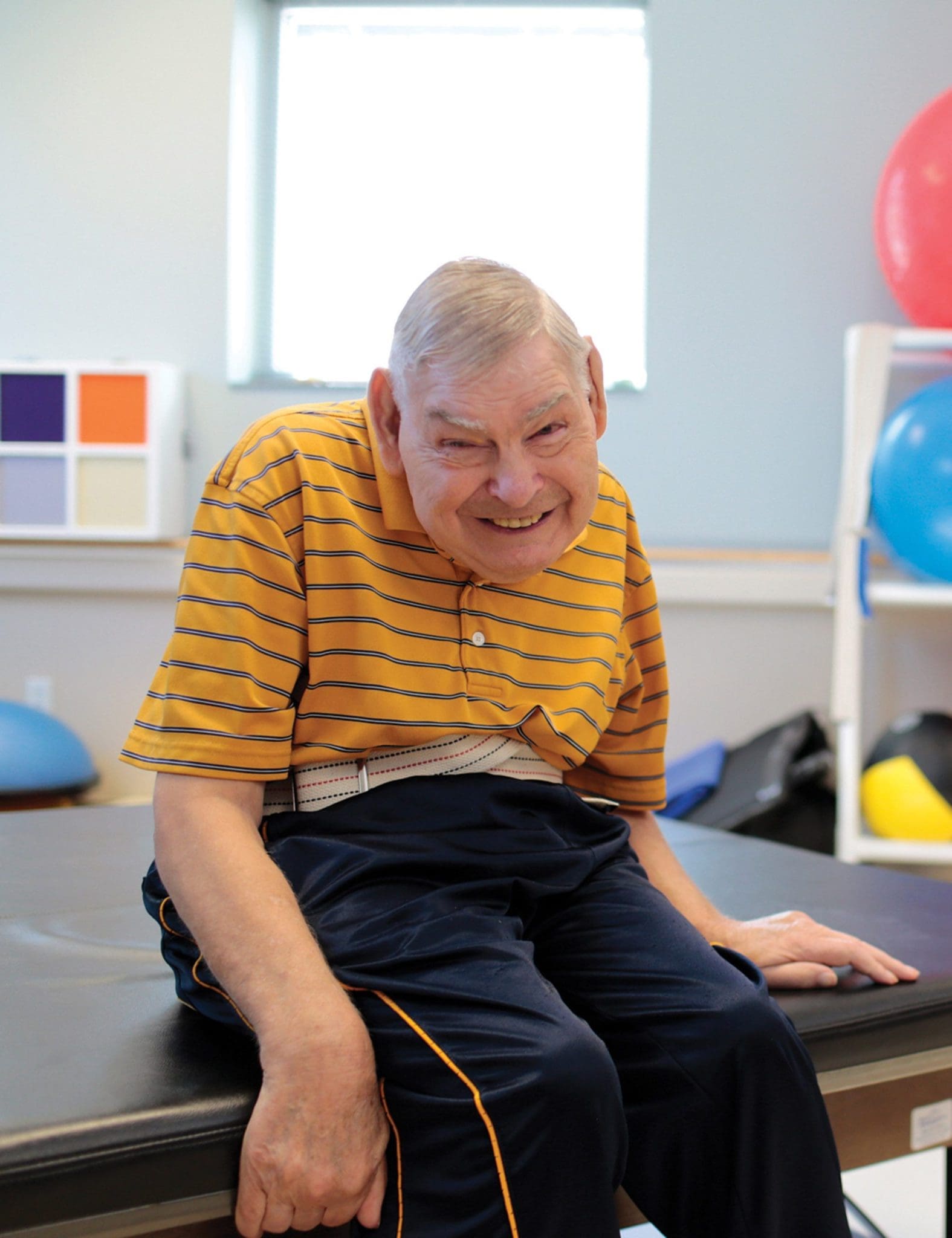 Smiling elderly man with an intellectual and developmental disability (IDD) sits on a therapy table, ready for his rehabilitation session to improve mobility and independence.